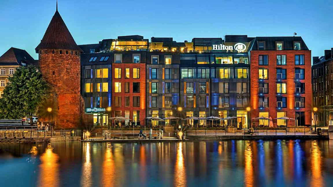 En historisk murbygning ved en moderne hotell ved vannet om kvelden. (AI-generert tekst)