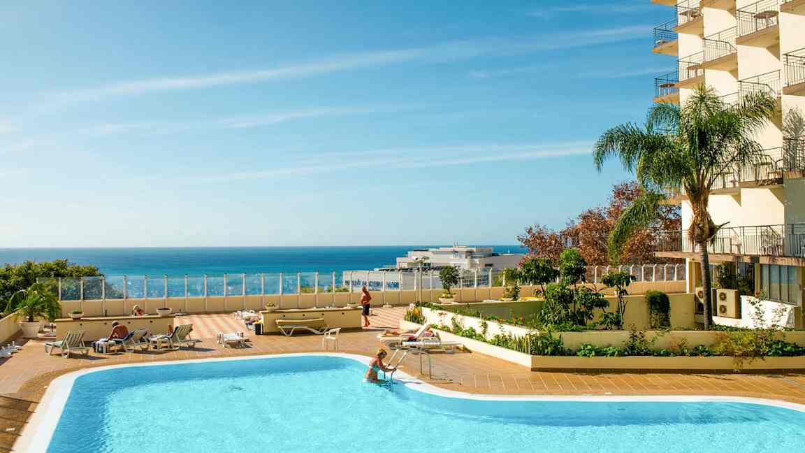 Et utendørs basseng med solsenger og en klar blå himmel over havet. (AI-generert tekst)