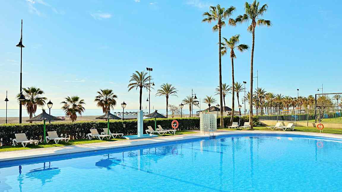 Et klart blått basseng med solsenger rundt, palmer i bakgrunnen og havet i det fjerne. (AI-generert tekst)