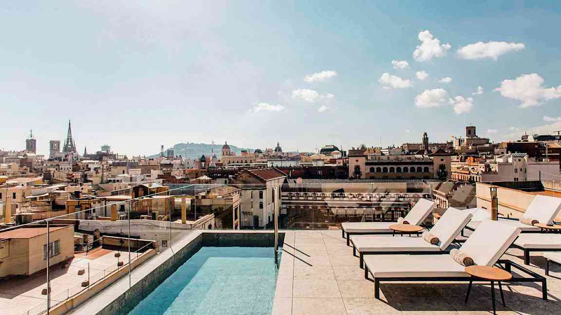 Et utendørsbasseng og solsenger på en takterrasse med utsikt over en by og dens bygninger under en klar himmel. (AI-generert tekst)
