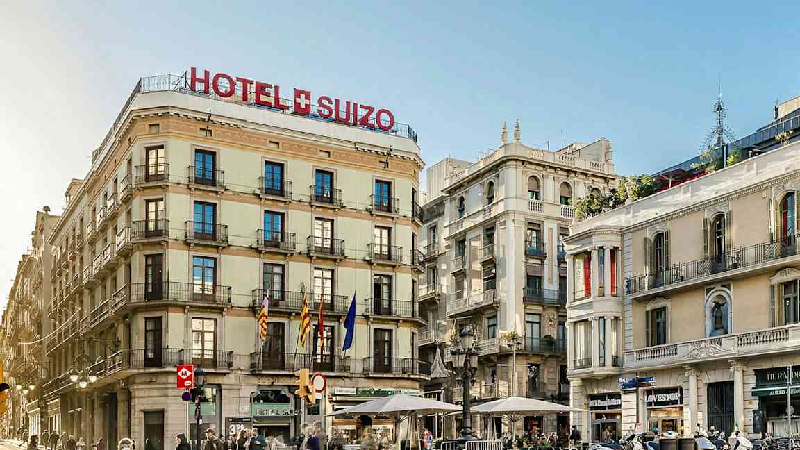 Et hotell kalt "Hotel Suizo" på et hjørne med flere etasjer og balkonger, omgitt av andre bygninger og mennesker som går på gaten. (AI-generert tekst)