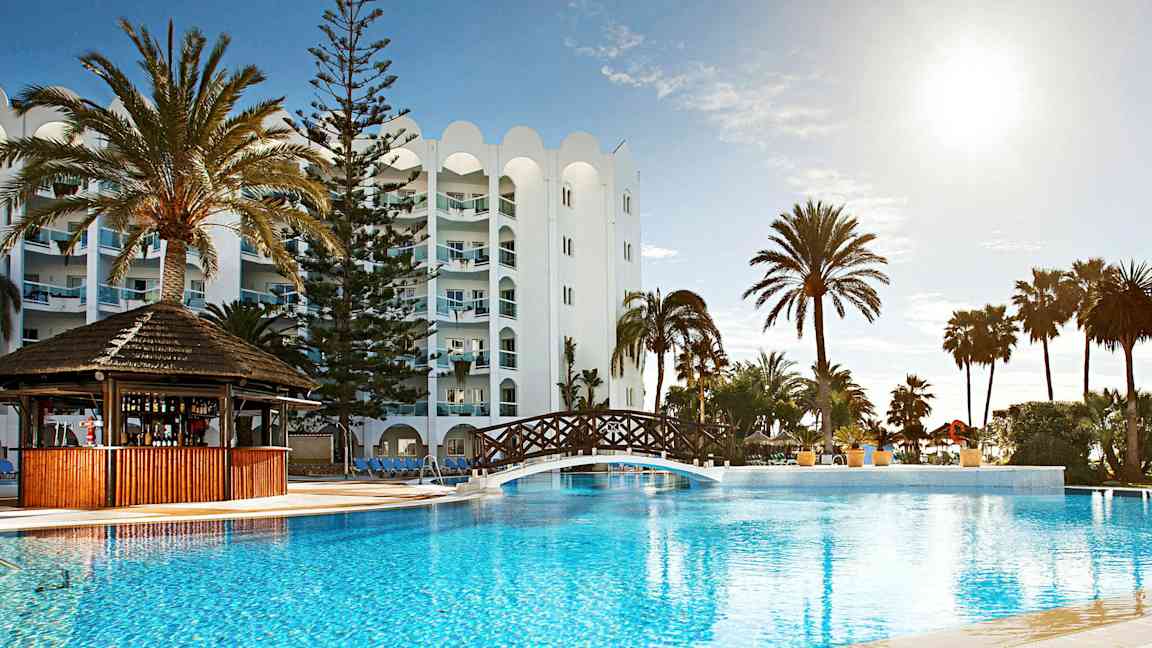 Bilde av et feriested med et utendørs basseng, en trebro over bassenget, palmer og en hvit bygning i bakgrunnen. (AI-generert tekst)