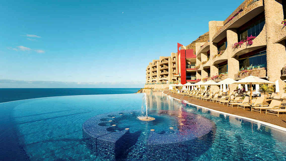 Et infinitybasseng ved strandkanten med solsenger og hotellbygninger i bakgrunnen. (AI-generert tekst)