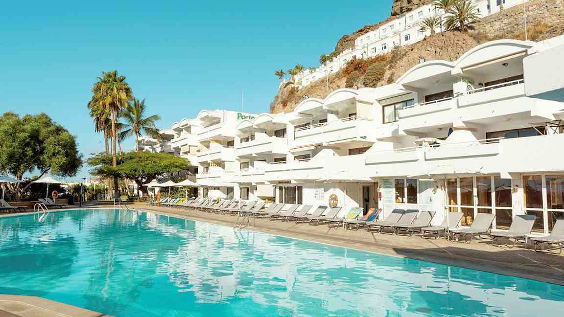 Et basseng foran et hvitt hotellkompleks med flere balkonger. I bakgrunnen vises palmer og en skråning. (AI-generert tekst)