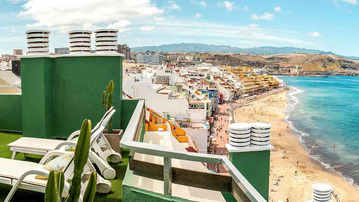 Utsikt fra en takterrasse med solsenger, kaktuser og en strand samt byen i bakgrunnen. (AI-generert tekst)
