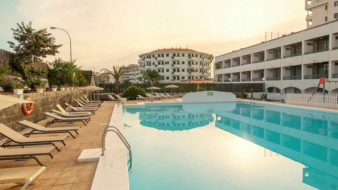 Utsikt over et basseng med solsenger ved siden av og omkringliggende bygninger. (AI-generert tekst)