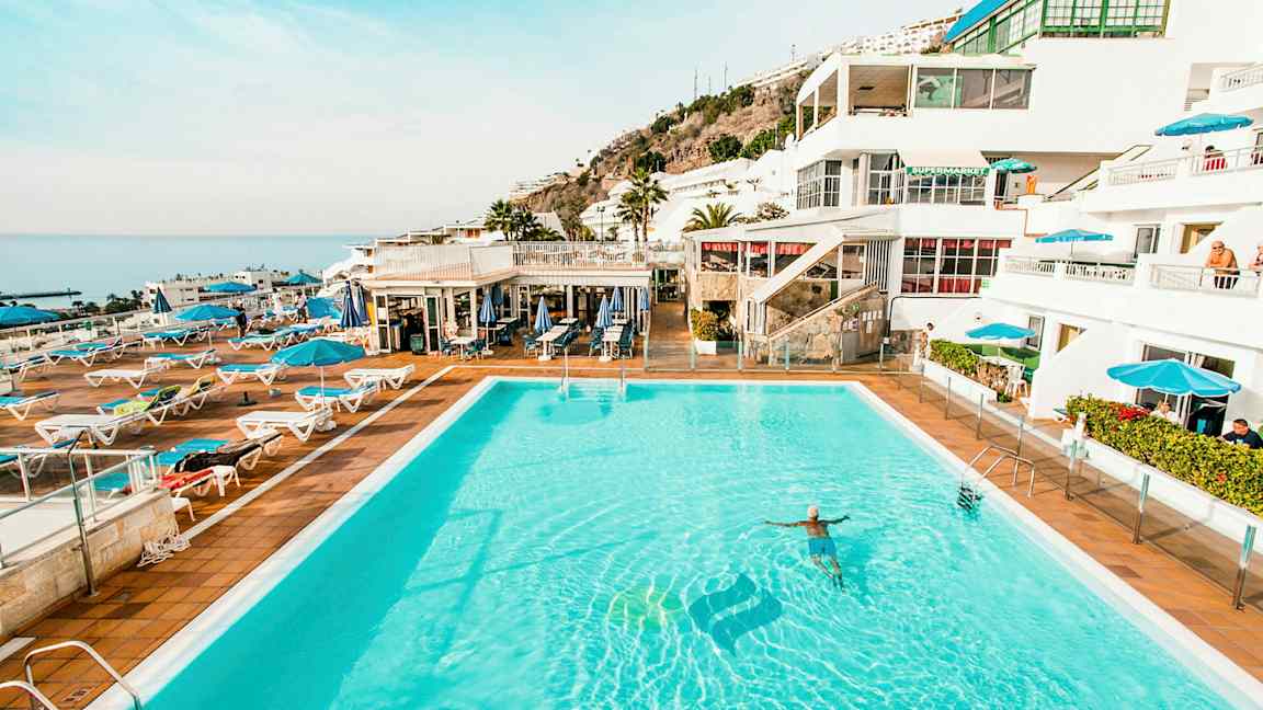 Bilde av et stort utendørs basseng omgitt av solsenger og parasoller med hvite bygninger og hav i bakgrunnen. (AI-generert tekst)