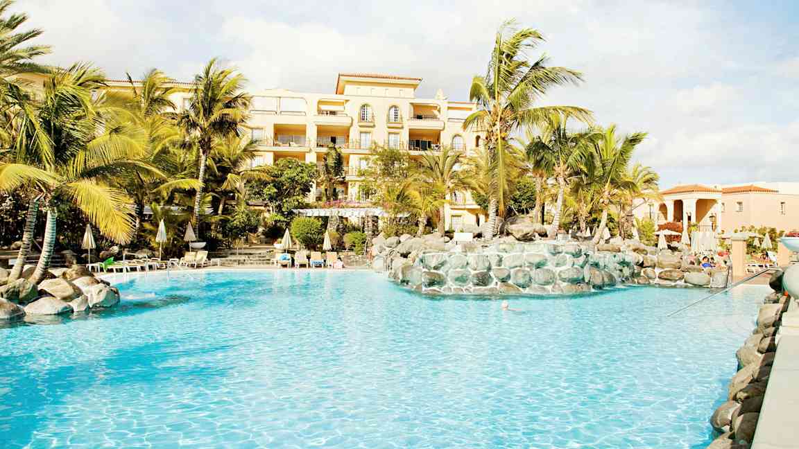 Bilde av et basseng med palmer og en bygning i bakgrunnen. (AI-generert tekst)