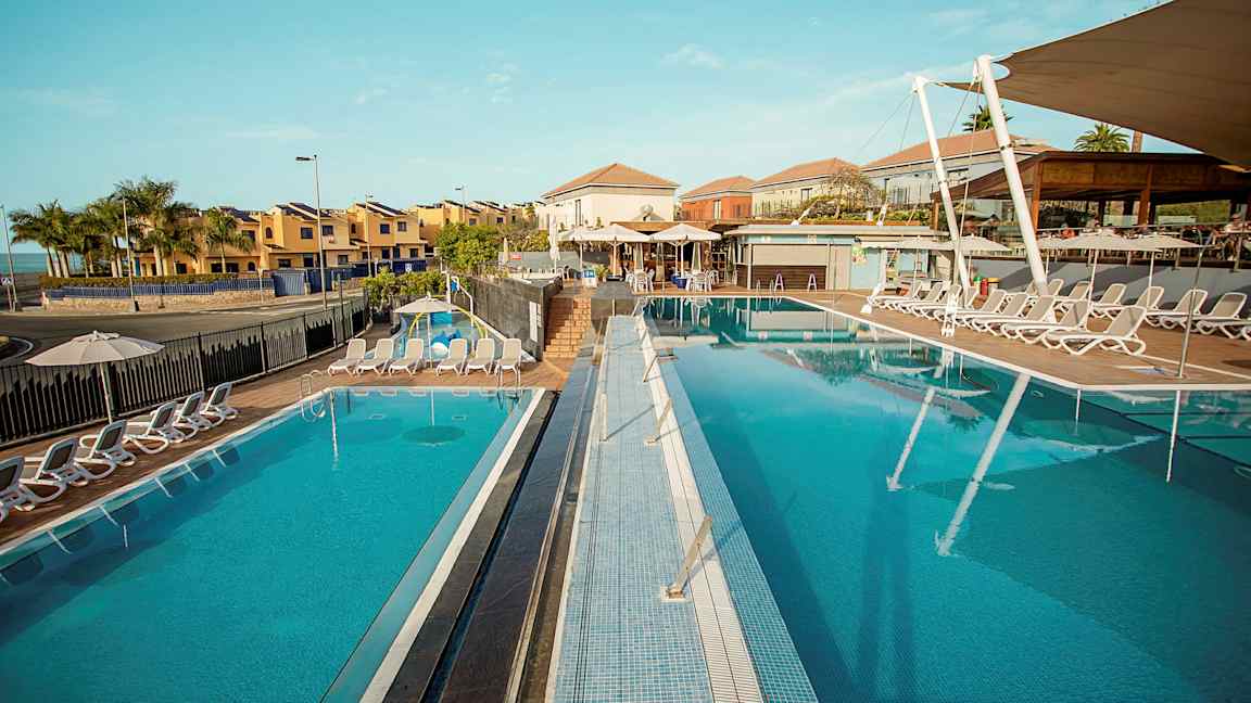 Et bilde av et solrikt feriested med to utendørsbassenger omgitt av solsenger, med bygninger i bakgrunnen. (AI-generert tekst)
