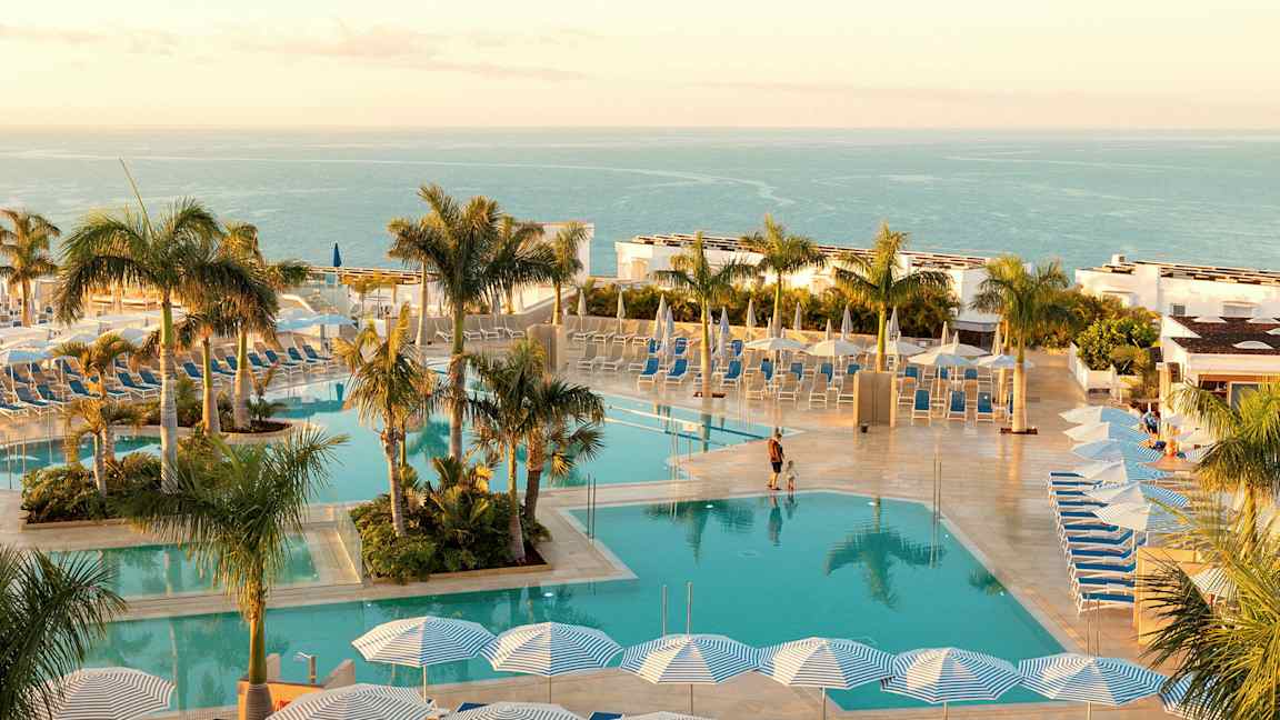 Et basseng med solsenger og parasoller, omgitt av palmer med havet i bakgrunnen. (AI-generert tekst)