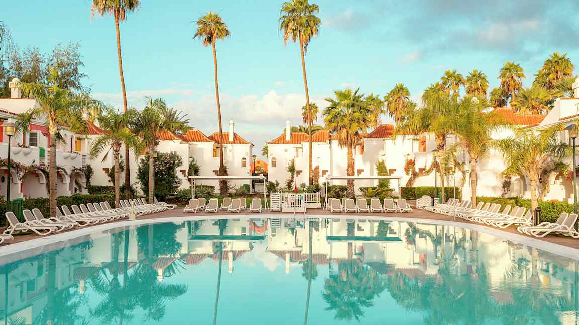 Hotellområde med hvitkalkede bygninger med røde tak, omgitt av palmer, og et stort basseng med solsenger rundt. (AI-generert tekst)