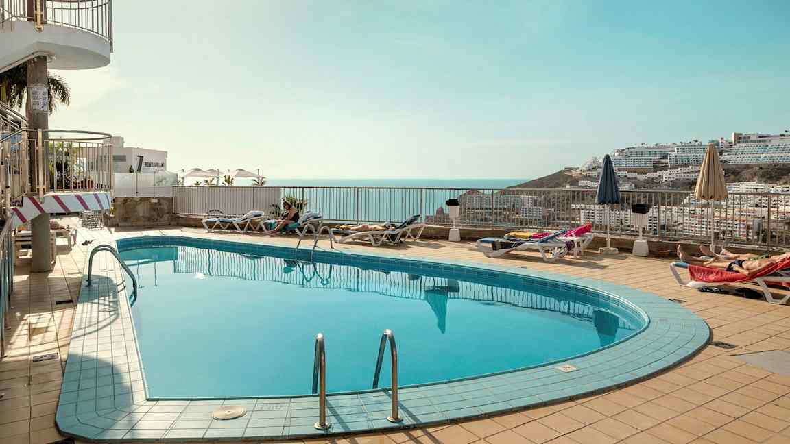 Et utendørs svømmebasseng med solsenger og parasoller rundt, med utsikt over havet og omkringliggende bygninger i bakgrunnen. (AI-generert tekst)