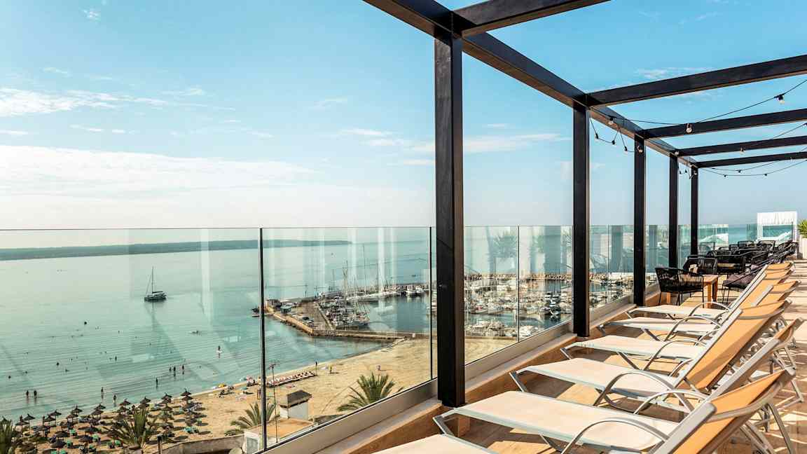 Solstoler satt opp foran glassrekkverk med utsikt over strand og havn. (AI-generert tekst)