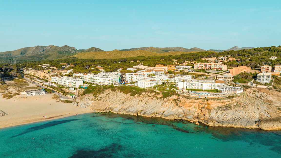 Strand med turkis vann foran en gruppe bolighus og hoteller ved en klipperik kyst. (AI-generert tekst)