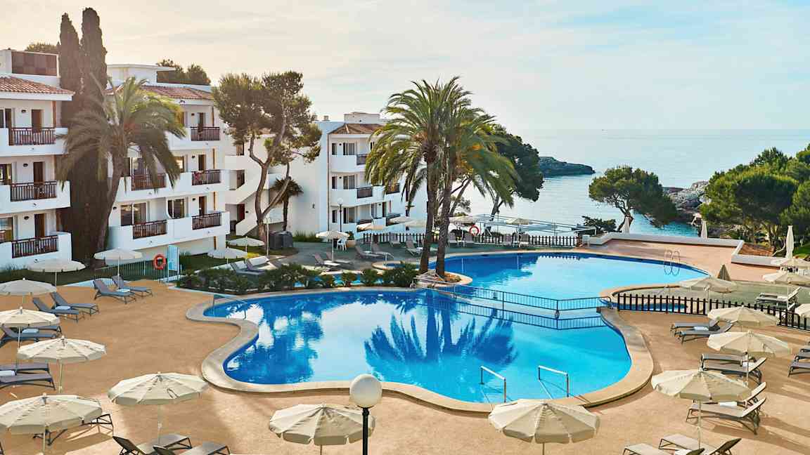 Bilde av et hotellområde med et stort basseng omgitt av solsenger og parasoller. I bakgrunnen sees havet og deler av bygningen. (AI-generert tekst)