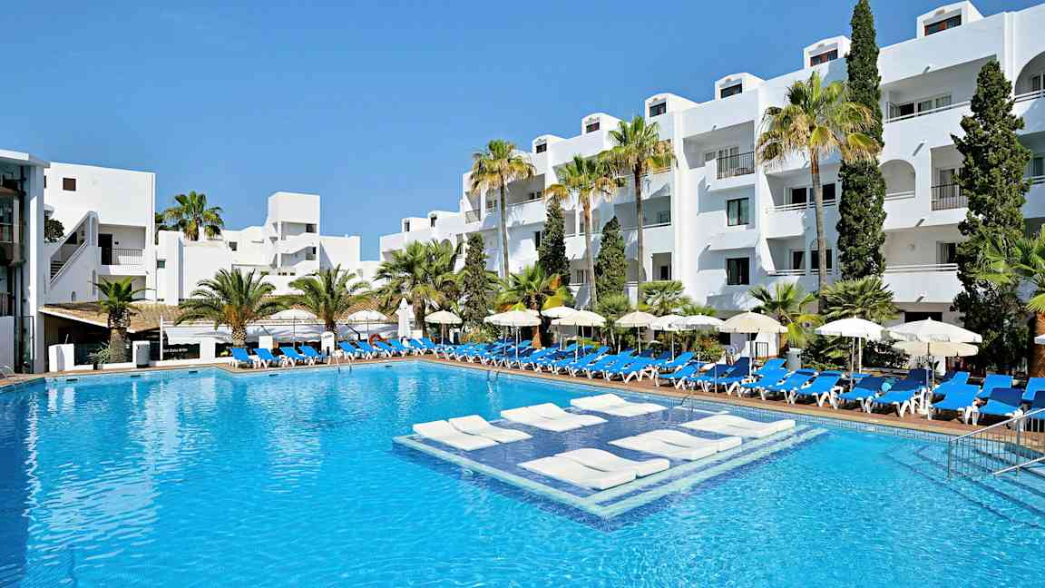 Et hotellbasseng med hvitmalte bygninger og palmer i bakgrunnen. (AI-generert tekst)