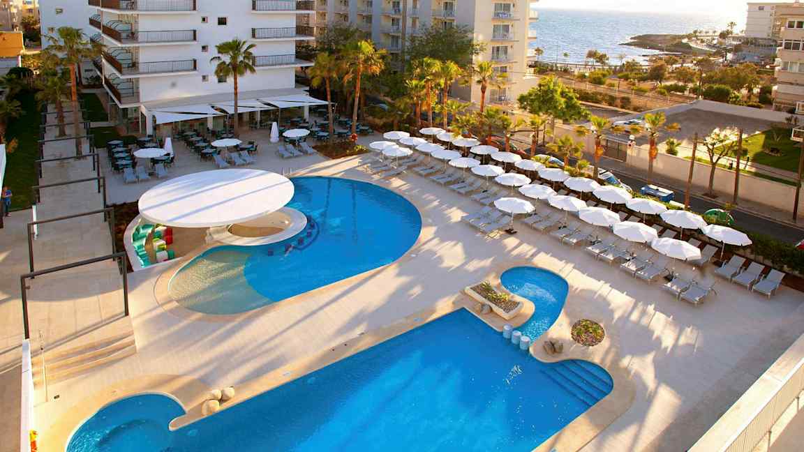 Hotellområde nær sjøen med basseng, solsenger og parasoller. (AI-generert tekst)