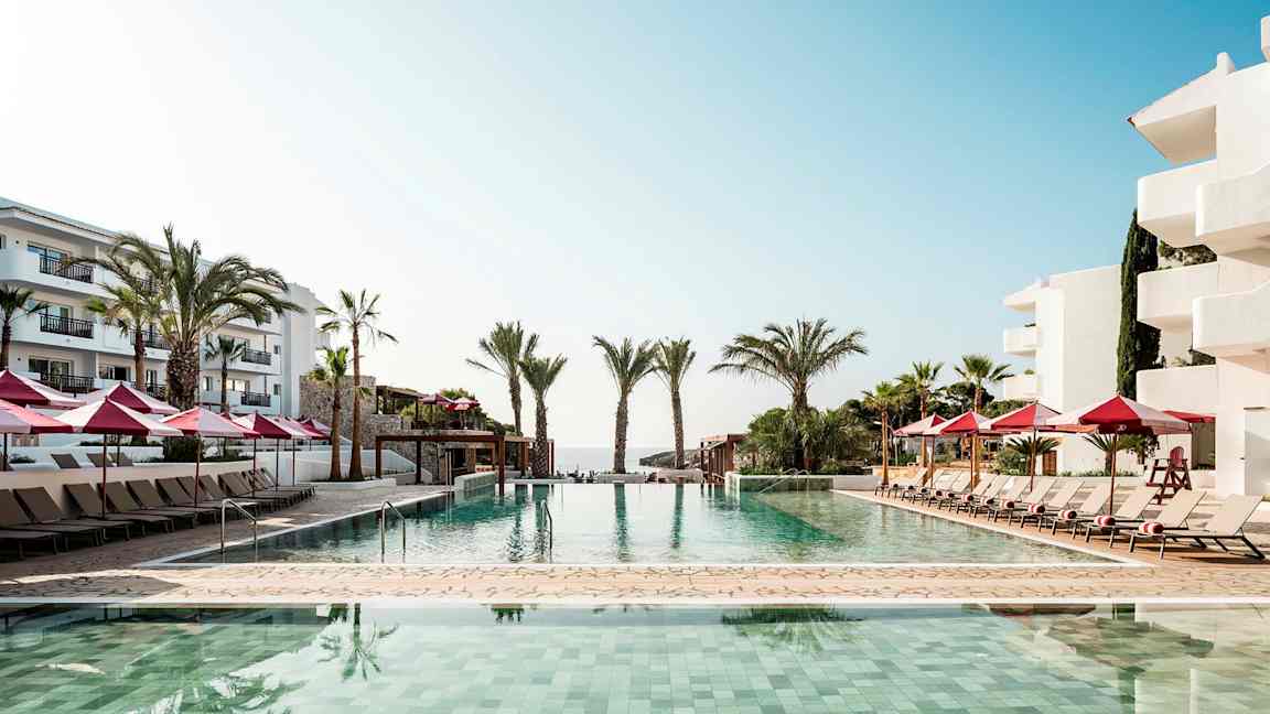 Utendørsbasseng ved kystnært hotell med solsenger og røde parasoller. (AI-generert tekst)