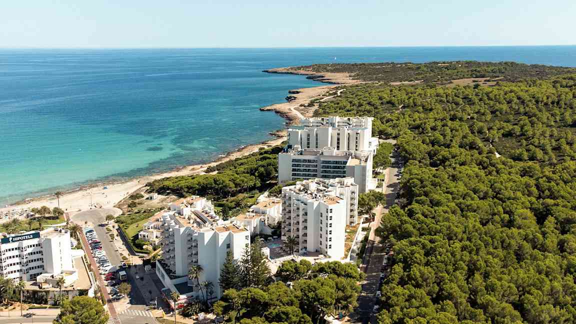 Luftfoto av en kystlinje med hotellbygninger foran en skog og nær en strand. (AI-generert tekst)