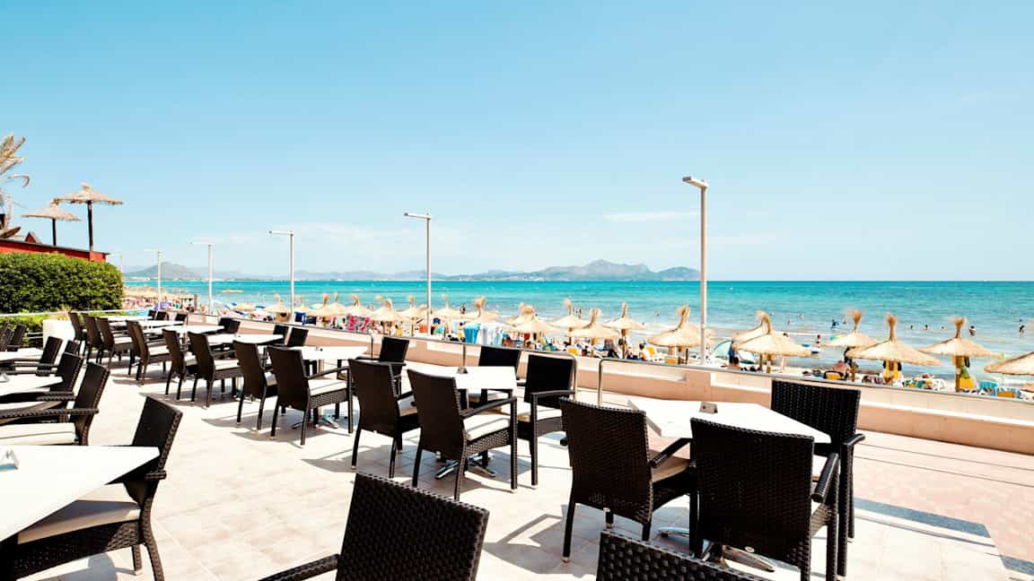 Uteservering med utsikt mot en sandstrand og havet med parasoller. (AI-generert tekst)