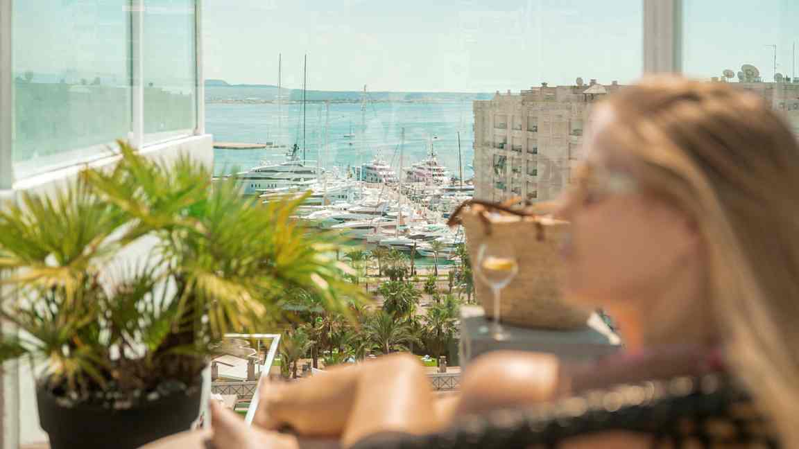 Utsikt mot båter i en marina, med en glassbalkong og frodige planter i forgrunnen. (AI-generert tekst)