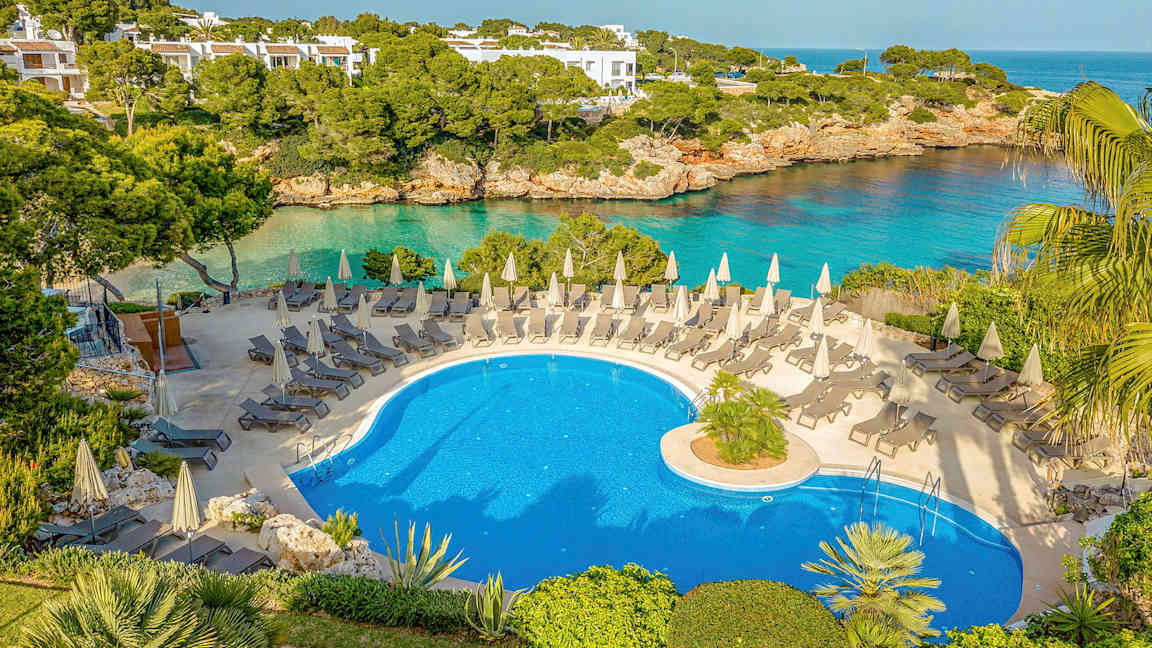 Et utendørs svømmebasseng med solsenger og parasoller, omgitt av grønne områder og med utsikt over havet. (AI-generert tekst)