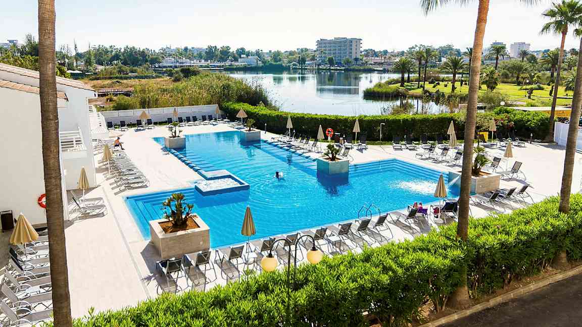 Utendørs svømmebasseng med solsenger og parasoller rundt, omgitt av grønne planter og med utsikt over en innsjø og et åpent landskap. (AI-generert tekst)