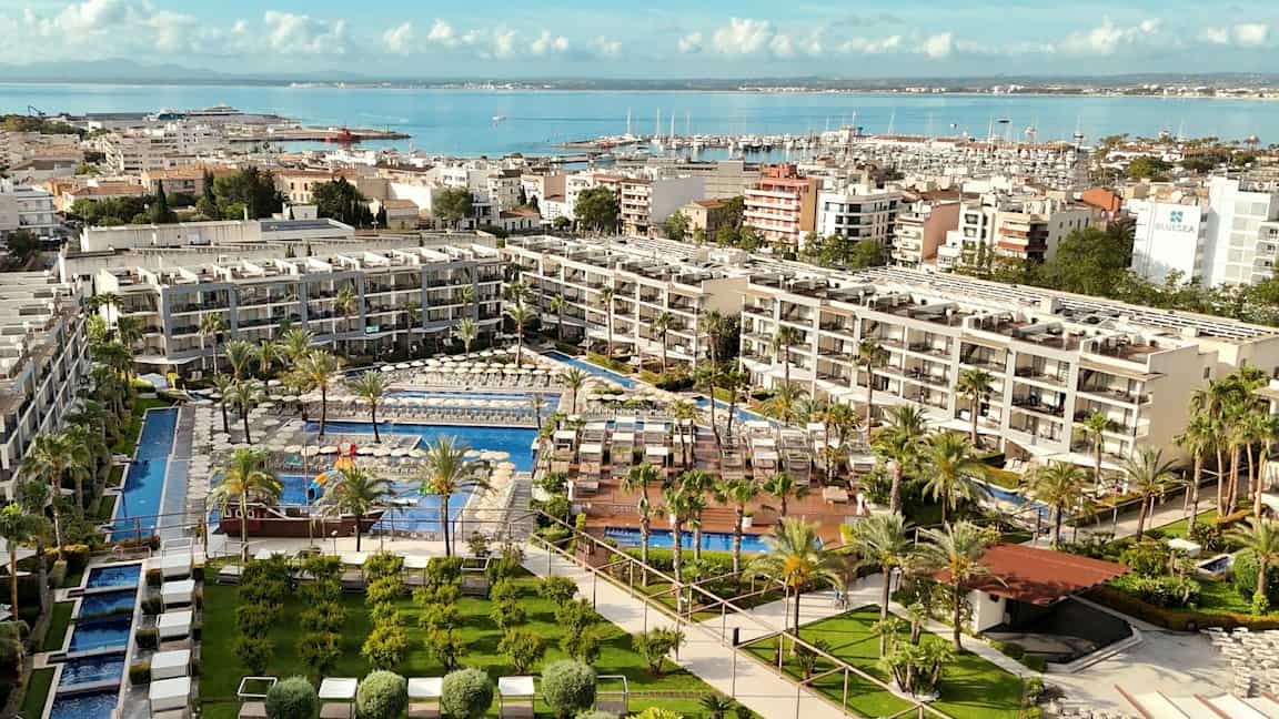 Et hotellkompleks med bassenger omgitt av grøntområder og solsenger, plassert ved vannet med en havn i bakgrunnen. (AI-generert tekst)