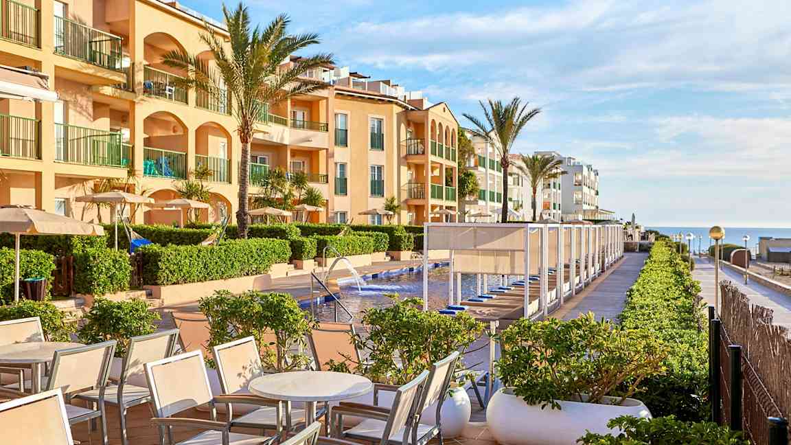 Et bilde av et hotellbasseng med solsenger og parasoller, omgitt av bygninger og palmer. (AI-generert tekst)