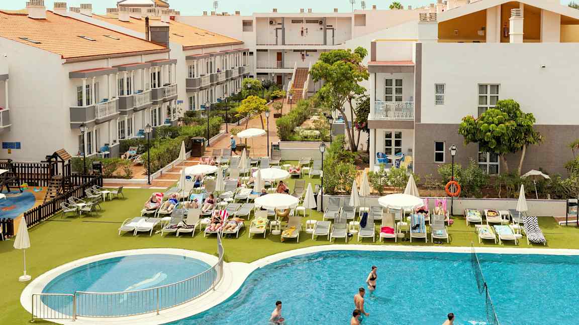 Hotellområde med bygninger, solsenger og et basseng. (AI-generert tekst)