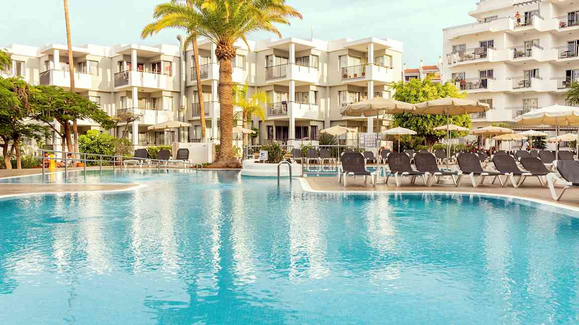 Utendørsbasseng omgitt av solsenger og parasoller, med en moderne bygning i bakgrunnen. (AI-generert tekst)