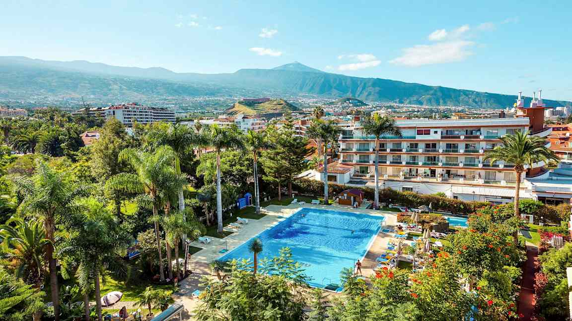 Et basseng omgitt av planter og bygninger med et fjell i bakgrunnen. (AI-generert tekst)