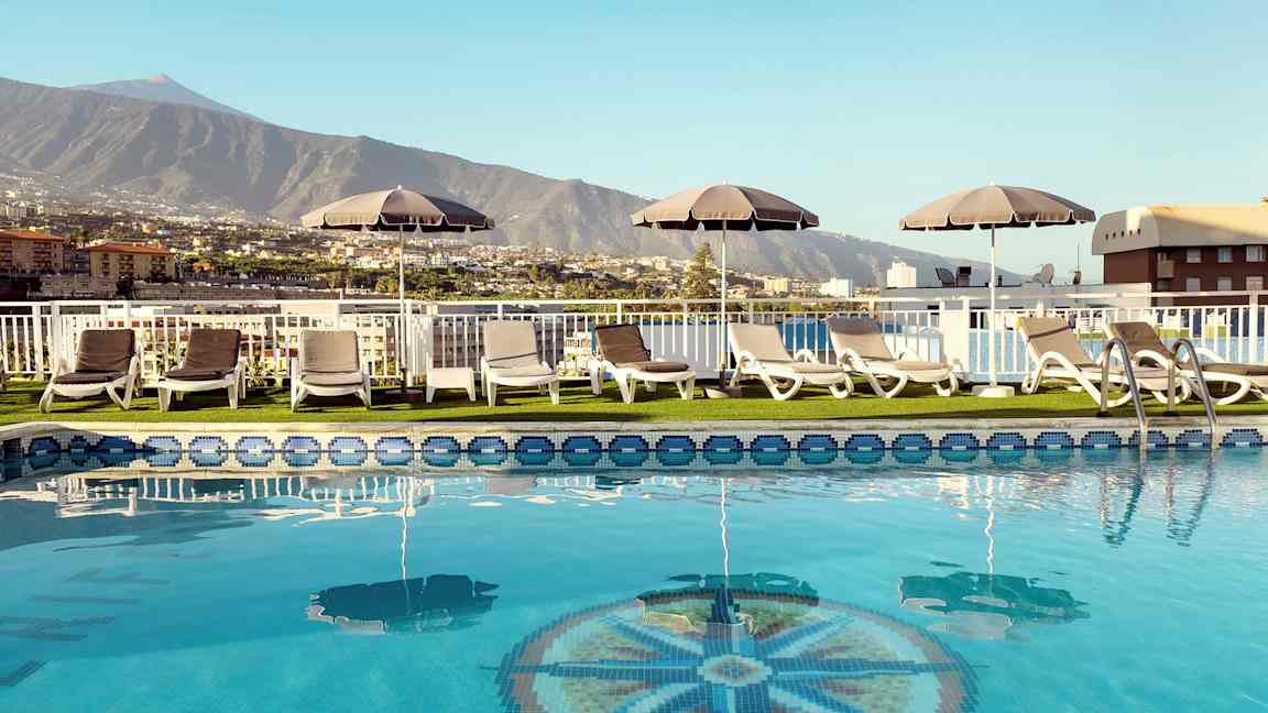 Bassengområde med solsenger og parasoller med utsikt over fjell og bygninger. (AI-generert tekst)
