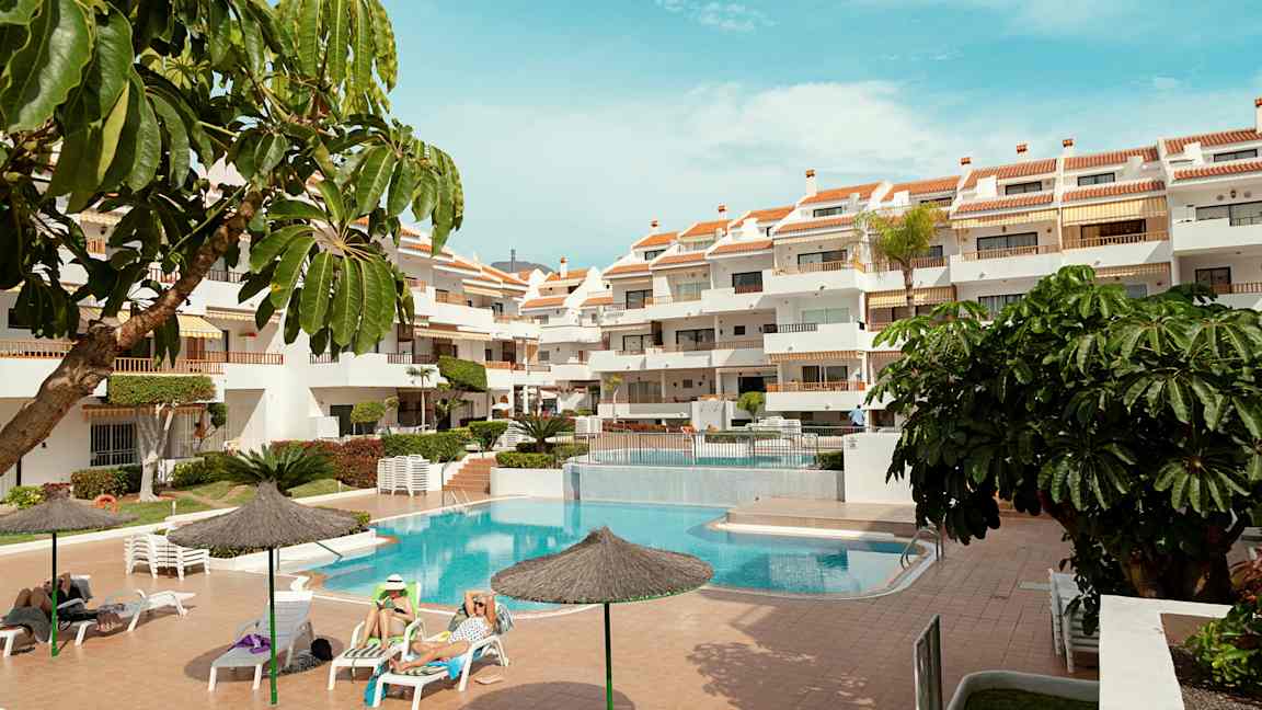 Et utendørsområde med et basseng, solsenger og et fleretasjehotell. Flere parasoller og grønne planter omgir området. (AI-generert tekst)