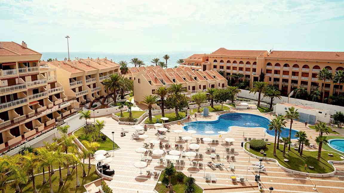 Flere bygninger med terrakottatak omgir et basseng og solsenger, med omkringliggende palmer og havet i bakgrunnen. (AI-generert tekst)