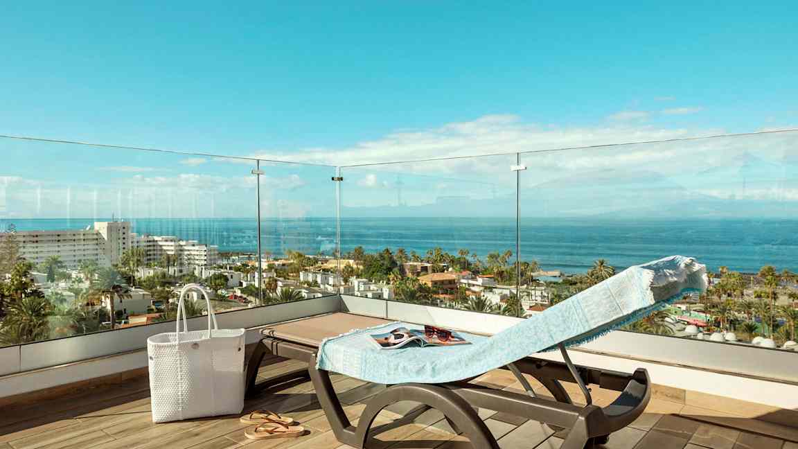 Utsikt fra en terrasse med en solseng dekket med et håndkle, en hvit veske og sandaler, over man ser havet og bygninger. (AI-generert tekst)