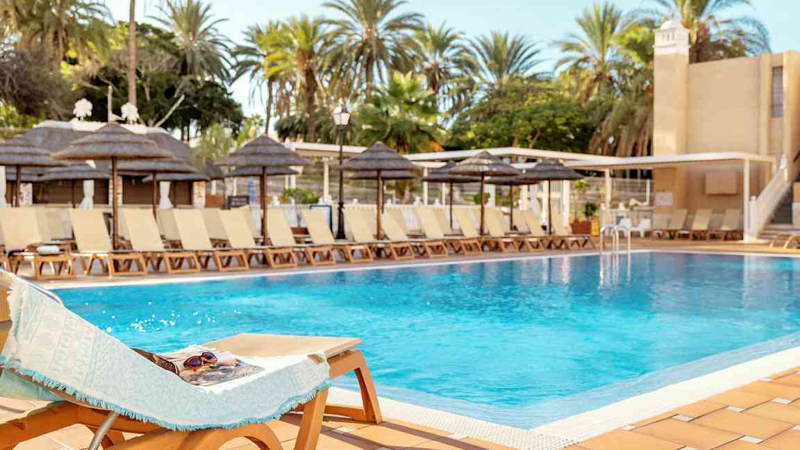 Et basseng omgitt av solsenger og palmer under en klar himmel. (AI-generert tekst)