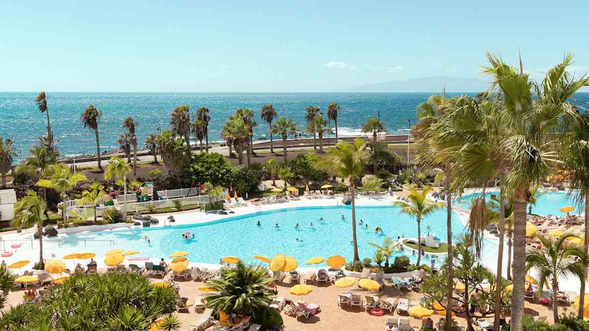 Bassengområde med solsenger, parasoller og mennesker, med havet i bakgrunnen. (AI-generert tekst)