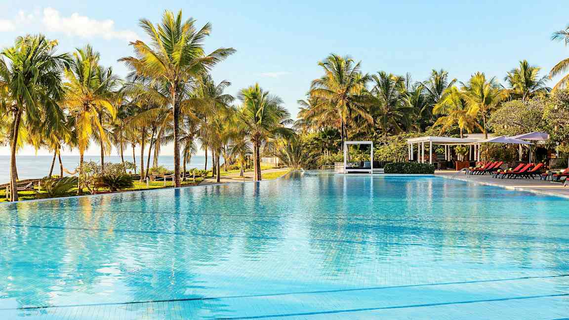 Et stort utendørsbasseng omgitt av palmer, med solsenger og paviljonger i bakgrunnen. (AI-generert tekst)