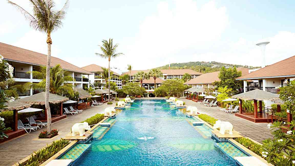 Et stort basseng på et feriested omgitt av palmer, solsenger og bungalowlignende bygninger. (AI-generert tekst)