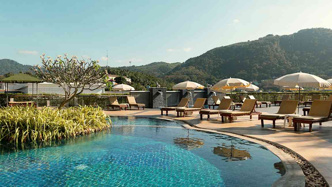 Utendørs basseng med solsenger og parasoller, omgitt av grøntområder og fjell i bakgrunnen. (AI-generert tekst)