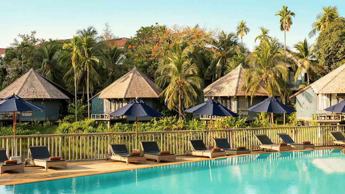 Solstoler og parasoller ved et svømmebasseng med bungalowlignende bygninger og vegetation i bakgrunnen. (AI-generert tekst)