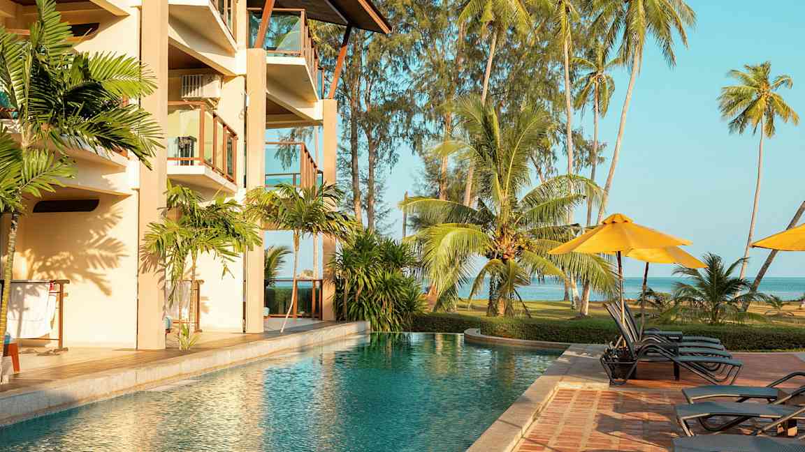 Et utendørs svømmebasseng ved et hotell med balkonger, omgitt av tropiske palmer og gressplen ved sjøen. (AI-generert tekst)