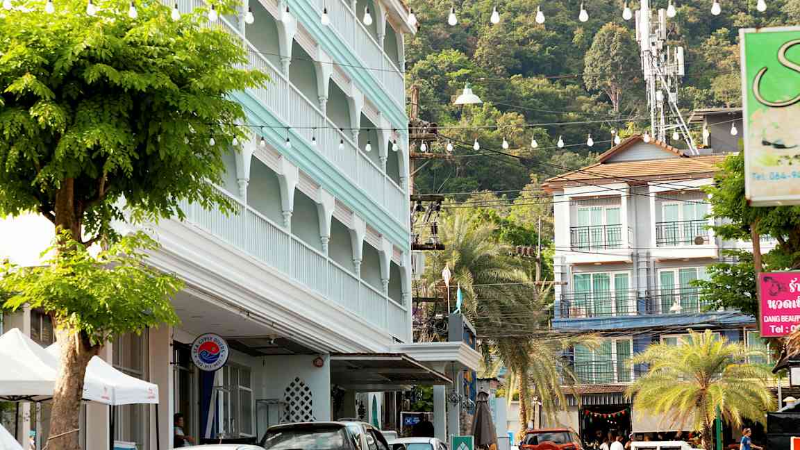 Gatebilde med parkerte biler, en bygning med blå fasade og frodige trær. (AI-generert tekst)