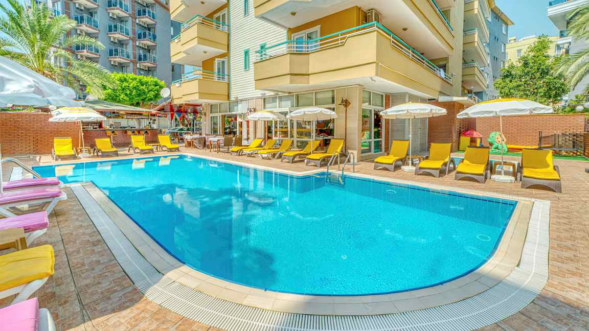 Et utendørs basseng omgitt av solsenger og parasoller foran en hotellbygning. (AI-generert tekst)