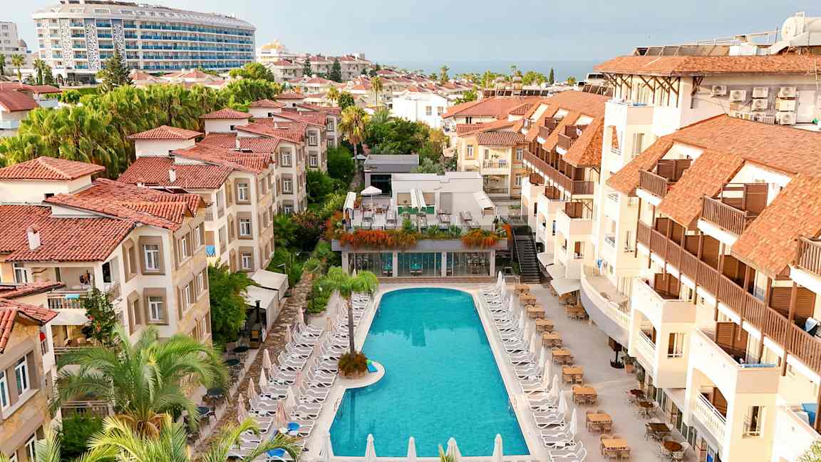 Hotellområde med et basseng omgitt av solsenger og bygninger med røde tak. (AI-generert tekst)