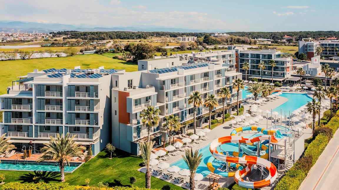 Flyfoto av et hotell med bassenger og vannsklier, omgitt av grøntområder. (AI-generert tekst)