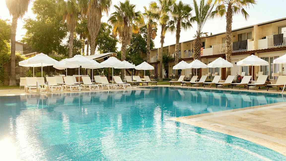 Basseng med solsenger og parasoller, omgitt av palmer og bygninger i bakgrunnen. (AI-generert tekst)