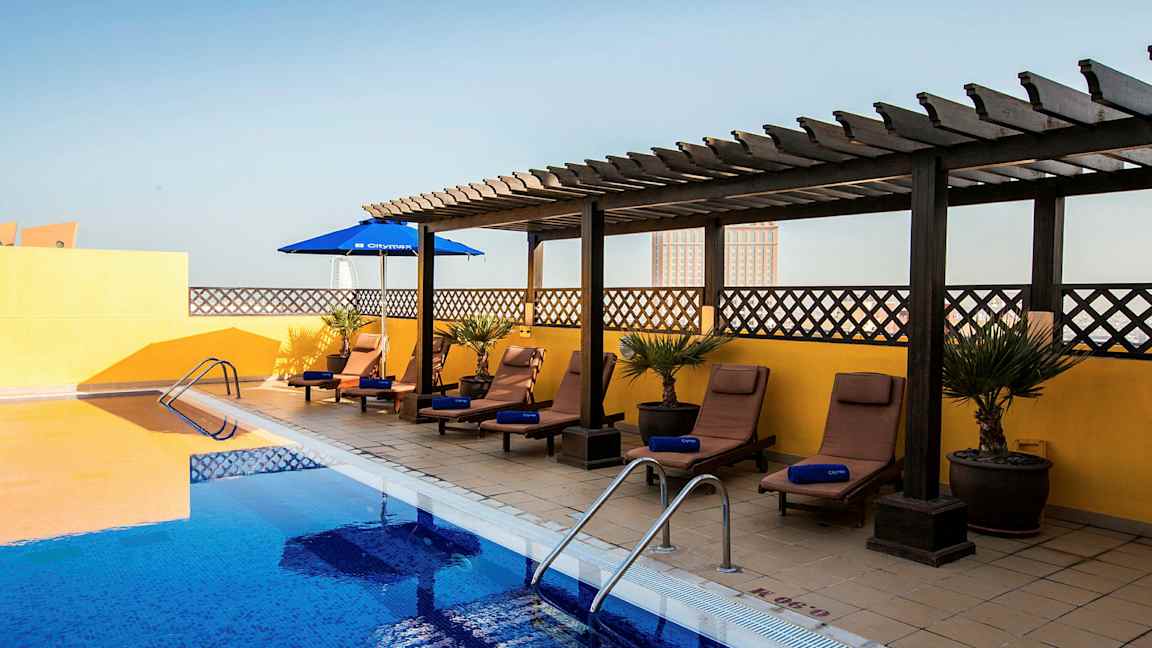 Et utendørsbasseng med blått vann omgitt av solsenger og parasoller. Solsengene står på et belagt gulv med en gul vegg og trepergola i bakgrunnen. (AI-generert tekst)