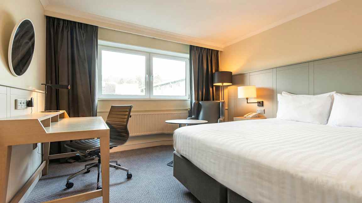 Bilde av et hotellrom med en seng, skrivebord og stol, og en lenestol ved siden av et vindu. (AI-generert tekst)